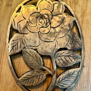 May include: A gold-toned metal trivet in the shape of a rose with leaves. The trivet is designed with an openwork pattern, allowing heat to escape.