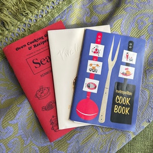 May include: Three vintage cookbooks with colorful covers. The first cookbook is red with the title "Oven Cooking & Recipes". The second cookbook is white with the title "Sea Tive". The third cookbook is blue with the title "metropolitan COOK BOOK".