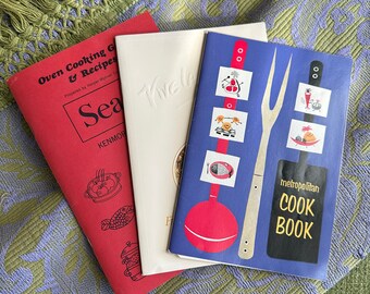 May include: Three vintage cookbooks with colorful covers. The first cookbook is red with the title "Oven Cooking & Recipes". The second cookbook is white with the title "Sea Tive". The third cookbook is blue with the title "metropolitan COOK BOOK".