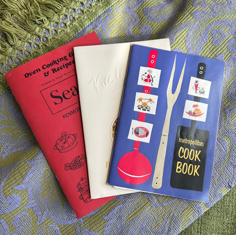 May include: Three vintage cookbooks with colorful covers. The first cookbook is red with the title "Oven Cooking & Recipes". The second cookbook is white with the title "Sea Tive". The third cookbook is blue with the title "metropolitan COOK BOOK".