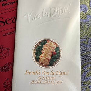 May include: A white cookbook with the title "Vive la Dijon!" in a bold, embossed font. The cover features a photo of a dish with chicken, bacon, and broccoli in a white and gold serving dish. The text "French's Vive la Dijon! Signature Recipe Collection" is printed below the photo.
