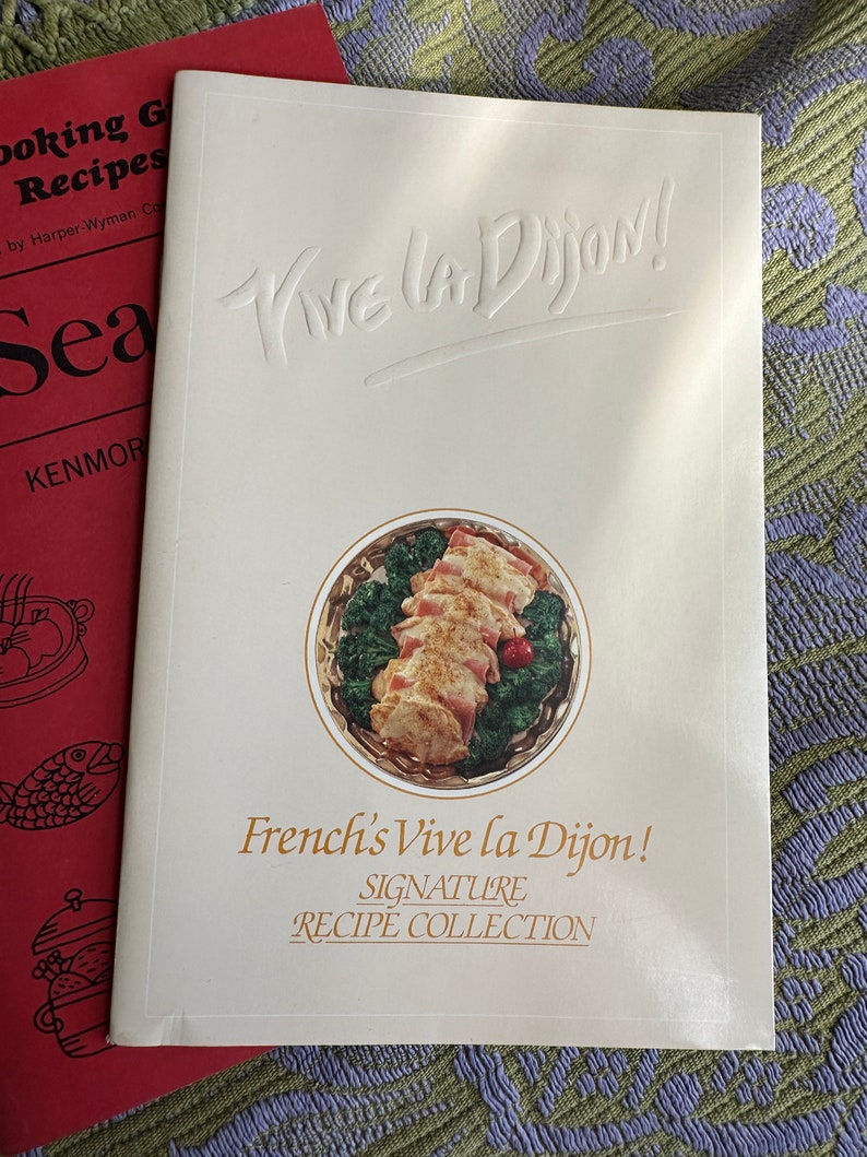 May include: A white cookbook with the title "Vive la Dijon!" in a bold, embossed font. The cover features a photo of a dish with chicken, bacon, and broccoli in a white and gold serving dish. The text "French's Vive la Dijon! Signature Recipe Collection" is printed below the photo.