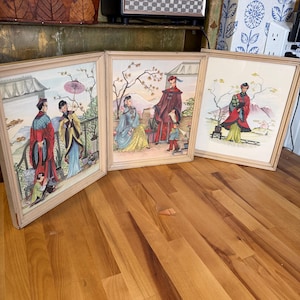 May include: Three framed vintage prints featuring Asian-inspired scenes. Each print depicts figures in traditional attire, with vibrant colors and detailed backgrounds. The frames are light-colored wood, adding a classic touch to the artwork. The prints are displayed on a wooden floor.