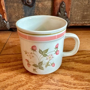 May include: White ceramic mug with a pink and green stripe design. The mug features a strawberry plant illustration with pink berries and green leaves. The mug has a curved handle.