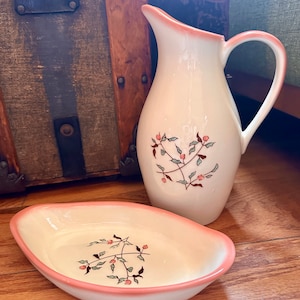 May include: A white ceramic pitcher and a white ceramic oval dish with a pink rim. Both pieces feature a floral design with green leaves and pink flowers.