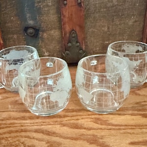 May include: Set of four clear glass mugs with a world map etched on the side. The mugs have a rounded shape and a handle.