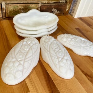 May include: A collection of white, vintage glass serving dishes. The set includes four small, heart-shaped dishes and three oval-shaped, footed dishes with intricate, embossed patterns. The dishes are arranged on a wooden surface.
