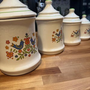 May include: A set of four white ceramic kitchen canisters with lids. Each canister features a colorful floral and bird design in orange, blue, and green. The lids have a green trim and a green knob. The canisters are arranged on a wooden surface.