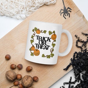 Può includere: Tazza in ceramica bianca con un design di Halloween. La tazza presenta un bordo di vite verde con zucche arancioni e il testo "Trick or Treat".