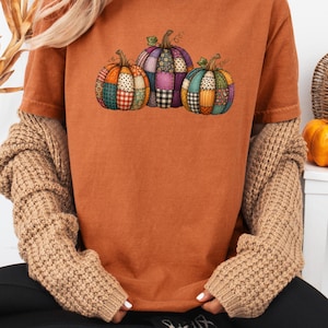 Peut inclure: Un t-shirt à manches courtes orange brûlé avec une impression graphique de trois citrouilles au motif patchwork coloré. Les citrouilles sont décorées de divers motifs, notamment des carreaux, des fleurs et des pois dans des tons orange, violet et bleu.