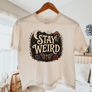 May include: A cream-colored short-sleeve t-shirt featuring a graphic print with the text STAY WEIRD. The design includes a crescent moon, a mushroom, a moth with a skull pattern, and various flowers surrounded by dark foliage and stars.