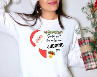 May include: White long-sleeve sweatshirt with a Santa Claus graphic on the front. The graphic includes the text "Santa isn't the only one JUDGING you". The Santa graphic is red and white. The sweatshirt is a festive holiday item.