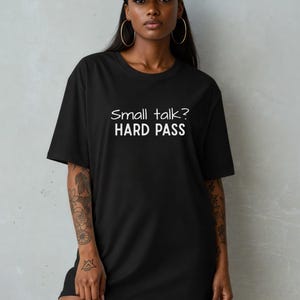 May include: Black t-shirt with the text "Small talk? HARD PASS" in white. The shirt is a casual, oversized style. The model is wearing a black cap and gold hoop earrings.
