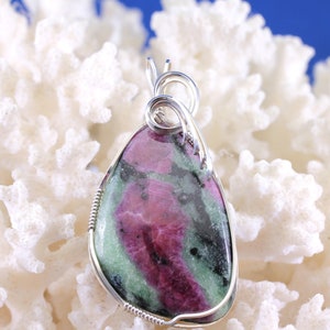 May include: A teardrop-shaped pendant featuring a polished ruby zoisite stone, wrapped in silver wire. The stone displays a mix of deep pink, green, and black hues. The pendant is displayed against a white coral-like background.
