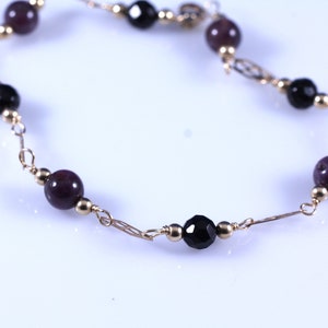 May include: A delicate gold-toned necklace featuring alternating dark purple and black beads. The necklace has small gold beads and decorative gold chain links. The beads are round and faceted.