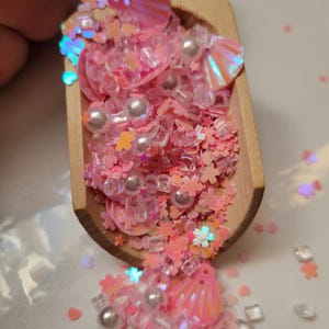 May include: A wooden scoop filled with pink and iridescent glitter, sequins, and beads. The glitter mix includes small stars, flowers, and seashells.