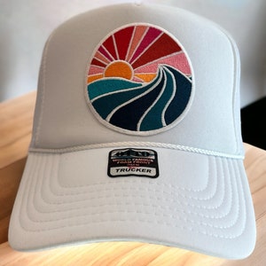 May include: White trucker hat with a colorful embroidered patch of a sunset over a wave. The patch is circular and has a red, orange, yellow, pink, blue, and green color scheme. The text "WORLD FAMOUS FOAM FRONT" and "TRUCKER" is on a black tag attached to the hat.