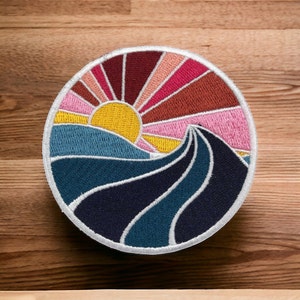 Peut inclure: Un patch brodé circulaire représentant un coucher de soleil coloré au-dessus d'une vague bleue et noire. Le soleil est jaune, le ciel est rose, orange et rouge, et la vague est soulignée de blanc.