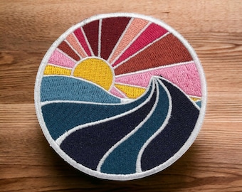 May include: A circular embroidered patch with a colorful sunset over a blue and black wave. The sun is yellow, the sky is pink, orange, and red, and the wave is outlined in white.