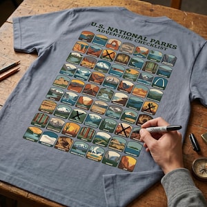 Pode incluir: Camiseta azul clara com um gráfico colorido dos Parques Nacionais dos EUA, projetada como uma lista de verificação. A camiseta exibe o texto "U.S. NATIONAL PARKS ADVENTURE CHECKLIST". Um marcador preto é usado para marcar os parques.