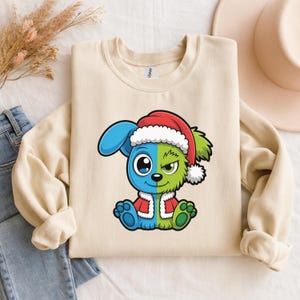 May include: A cream-colored sweatshirt featuring a cartoon character split in half, with blue and green sides. The character wears a red Santa hat with white trim and a matching red coat. The design is centered on the front of the sweatshirt.