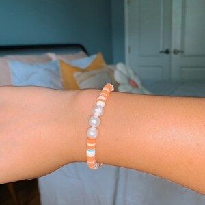 Op de afbeelding: Een armband met oranje en witte kralen, waaronder enkele parel-achtige kralen.
