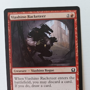 May include: A red, white, and black Magic: The Gathering card featuring a Viashino Racketeer creature with a 2/1 power and toughness. The card text reads: "When Viashino Racketeer enters the battlefield, you may discard a card. If you do, draw a card. "You may call me a fool, selling this stuff on Orzhov turf. Well, you need to be a little reckless to survive."
