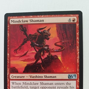 May include: A red and black Magic: The Gathering card featuring a red creature with horns and a tail. The card is titled "Mindclaw Shaman" and has a power and toughness of 2/2. The card text reads: "When Mindclaw Shaman enters the battlefield, target opponent reveals his or her hand. You may cast an instant or sorcery card from that player's hand without paying its mana cost. Nicol Bolas gives power to those who will abuse it most."