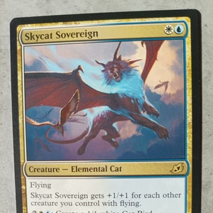 May include: A fantasy trading card featuring a white and red Skycat Sovereign creature with blue eyes and wings. The card text reads "Skycat Sovereign gets +1/+1 for each other creature you control with flying. 2: Create a 1/1 white Cat Bird creature token with flying. The bounty on its head is as old as Skysail, but no one has ever tried to collect."