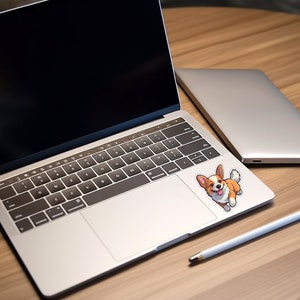 Pode incluir: Um computador port&aacute;til prateado com uma tela preta e um teclado branco. Um adesivo de um c&atilde;o corgi de desenho animado est&aacute; no touchpad. Um laptop prateado est&aacute; no fundo e uma caneta branca est&aacute; sobre a mesa de madeira.