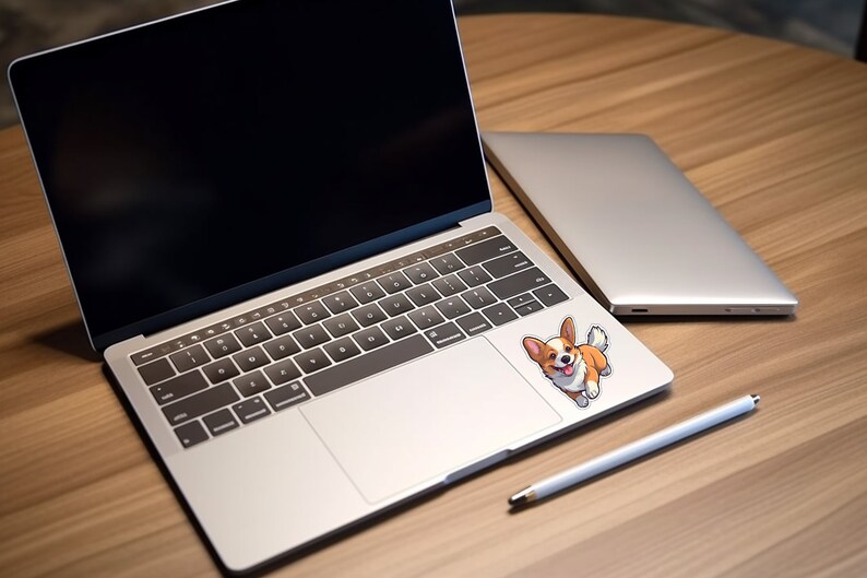 Pode incluir: Um computador port&aacute;til prateado com uma tela preta e um teclado branco. Um adesivo de um c&atilde;o corgi de desenho animado est&aacute; no touchpad. Um laptop prateado est&aacute; no fundo e uma caneta branca est&aacute; sobre a mesa de madeira.