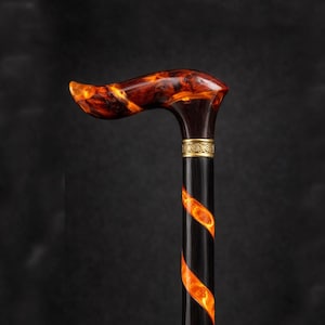 Könnte beinhalten: Dekorative Gehstock mit gebogenem Griff. Der Griff ist marmoriert orange und braun. Der Schaft ist schwarz mit einem leuchtenden orangefarbenen Spiraldesign. Am oberen Ende des Schafts befindet sich ein goldenes Band.