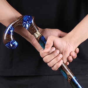May include: A wooden walking stick with a curved handle. The handle and upper shaft feature a swirling blue resin design, complemented by a brown wood grain. A silver-coloured metal band joins the handle to the shaft. The stick is held by a person.