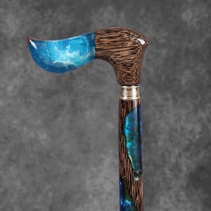 May include: A handcrafted walking cane with a curved handle. The handle features a blue resin design with white swirls, transitioning to a dark wood grain. The shaft incorporates blue and green resin accents, and is finished with a decorative metal band.
