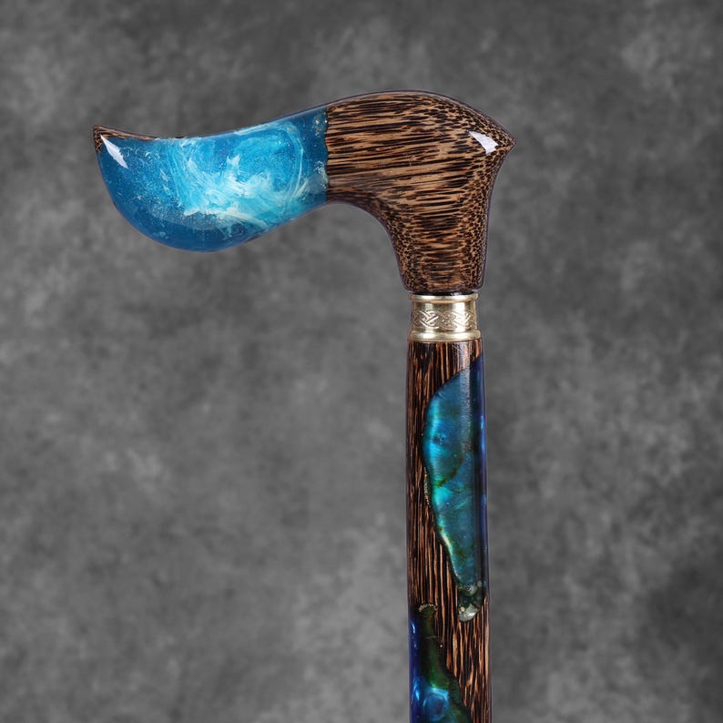May include: A handcrafted walking cane with a curved handle. The handle features a blue resin design with white swirls, transitioning to a dark wood grain. The shaft incorporates blue and green resin accents, and is finished with a decorative metal band.