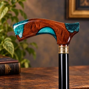 May include: A walking cane featuring a curved handle crafted from polished brown wood with inlaid teal resin accents. The handle connects to a smooth black shaft via a decorative gold-toned metal collar with an intricate embossed floral pattern.