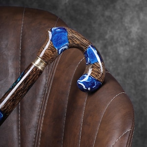 May include: A wooden walking cane with a curved handle. The handle features blue resin accents and a gold-colored metal band. The shaft is a dark brown color. The cane is resting on a brown leather chair.