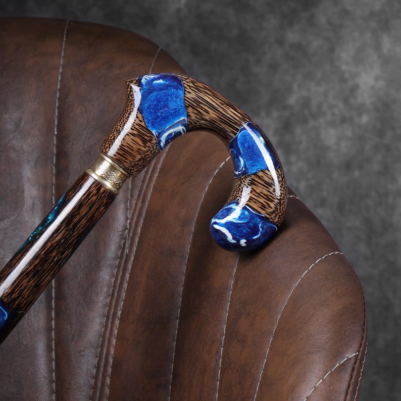 May include: A wooden walking cane with a curved handle. The handle features blue resin accents and a gold-colored metal band. The shaft is a dark brown color. The cane is resting on a brown leather chair.