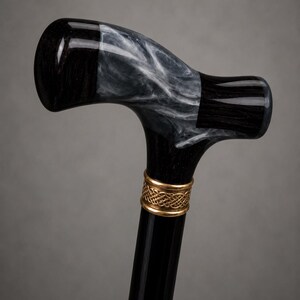 May include: A walking cane featuring a T-shaped handle made of black material with a swirling white marble-like pattern. A gold-colored metal collar with an intricate woven or Celtic-style knot design connects the handle to the smooth black shaft.
