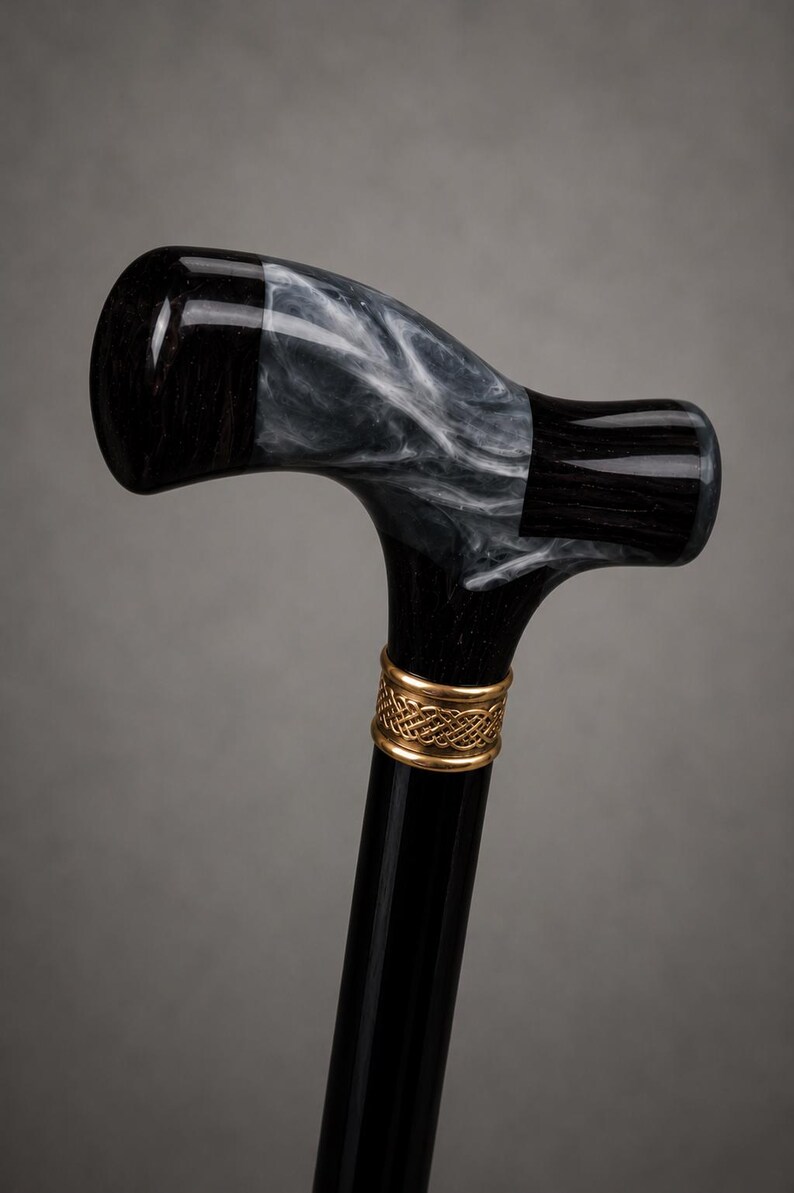 May include: A walking cane featuring a T-shaped handle made of black material with a swirling white marble-like pattern. A gold-colored metal collar with an intricate woven or Celtic-style knot design connects the handle to the smooth black shaft.