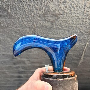 Puede incluir: Un mango de bast&oacute;n de resina azul y brillante con un dise&ntilde;o curvo. El mango est&aacute; montado sobre una base de madera marr&oacute;n oscuro con un anillo plateado. El mango es sostenido por una persona.