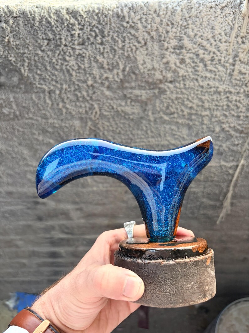 Puede incluir: Un mango de bast&oacute;n de resina azul y brillante con un dise&ntilde;o curvo. El mango est&aacute; montado sobre una base de madera marr&oacute;n oscuro con un anillo plateado. El mango es sostenido por una persona.