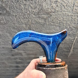 Puede incluir: Un mango decorativo, posiblemente para un bast&oacute;n o un bast&oacute;n, con un dise&ntilde;o de purpurina azul. El mango tiene una forma curva, con una base azul oscuro y una parte superior m&aacute;s clara llena de purpurina. Se asienta sobre una base oscura y redonda.
