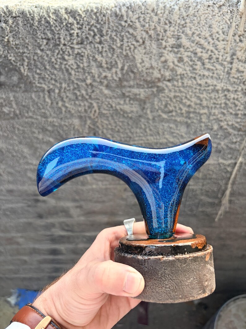 Puede incluir: Un mango decorativo, posiblemente para un bast&oacute;n o un bast&oacute;n, con un dise&ntilde;o de purpurina azul. El mango tiene una forma curva, con una base azul oscuro y una parte superior m&aacute;s clara llena de purpurina. Se asienta sobre una base oscura y redonda.