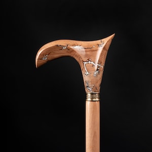 Peut inclure: Une canne en bois avec une poignée incurvée. La poignée est marron clair avec un motif décoratif d'accents argentés et marron foncé. Une bande dorée sépare la poignée de la tige marron clair. La canne est posée sur un fond noir.