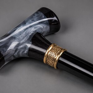 May include: A walking cane featuring a T-shaped handle with a black and white marbled resin finish. The handle connects to a smooth, glossy black shaft via a decorative gold-toned metal collar that displays an intricate, woven, Celtic-style pattern.