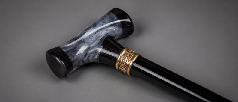 May include: A walking cane featuring a T-shaped handle with a black and white marbled resin finish. The handle connects to a smooth, glossy black shaft via a decorative gold-toned metal collar that displays an intricate, woven, Celtic-style pattern.