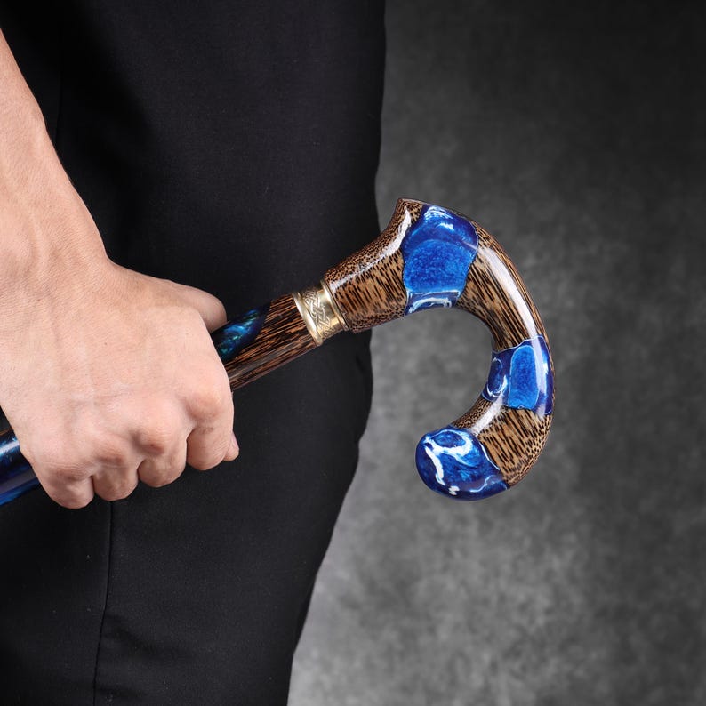 May include: A wooden cane with a curved handle, featuring a brown and blue design. The handle has a brass accent and is held by a person wearing a black shirt. The cane's design incorporates blue resin inlays.