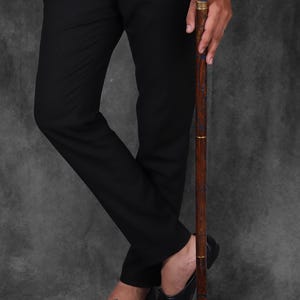 May include: A wooden walking stick with a blue resin inlay and a gold band. The stick has a curved handle and is being held by a person wearing black trousers and shoes.