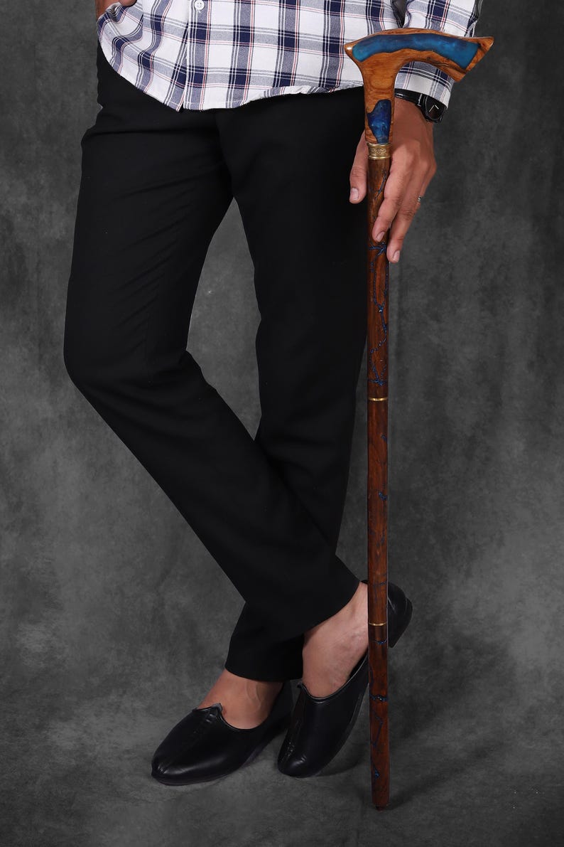 May include: A wooden walking stick with a blue resin inlay and a gold band. The stick has a curved handle and is being held by a person wearing black trousers and shoes.